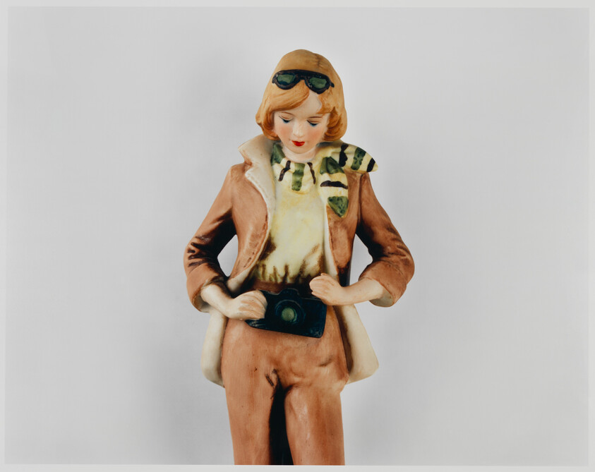 A ceramic figurine of a woman wearing goggles and a scarf while holding a camera.