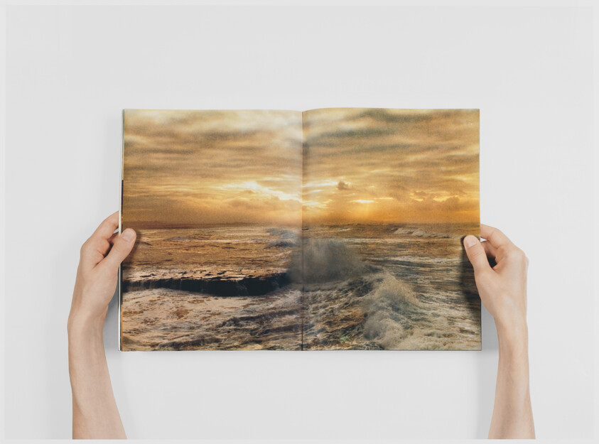 A person is holding a magazine open to a spread featuring a vibrant ocean sunset scene with waves crashing against rocks.