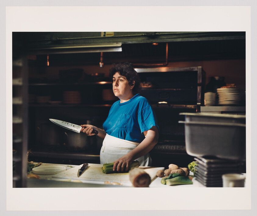 A cook in a blue shirt holds a large knife while preparing leeks at a kitchen counter.