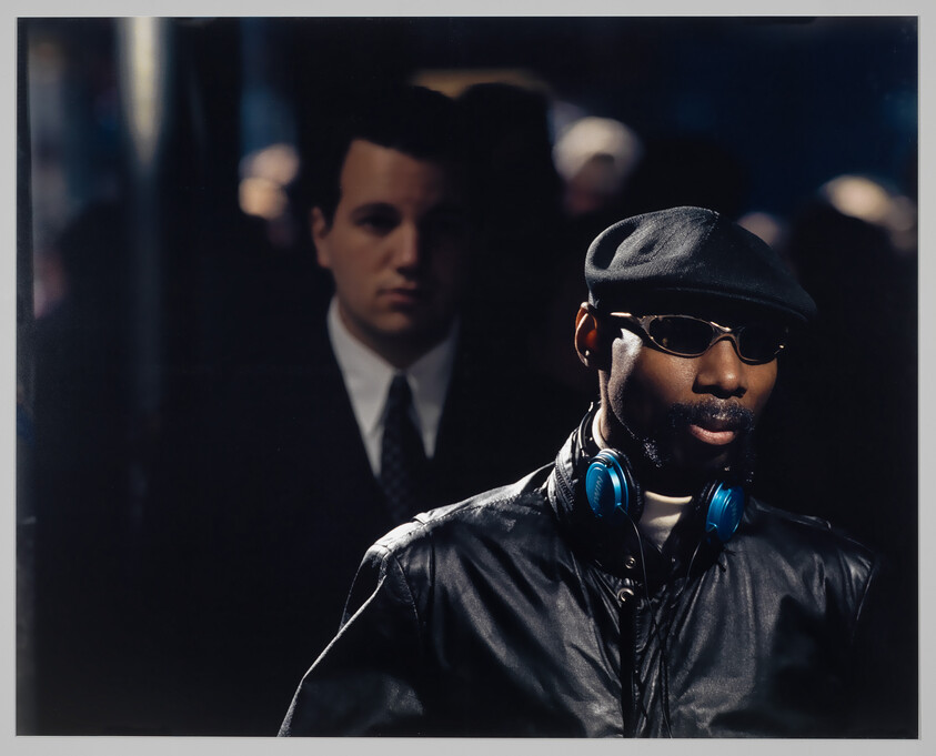 Man in leather jacket and cap wears blue headphones and sunglasses while walking in crowd.