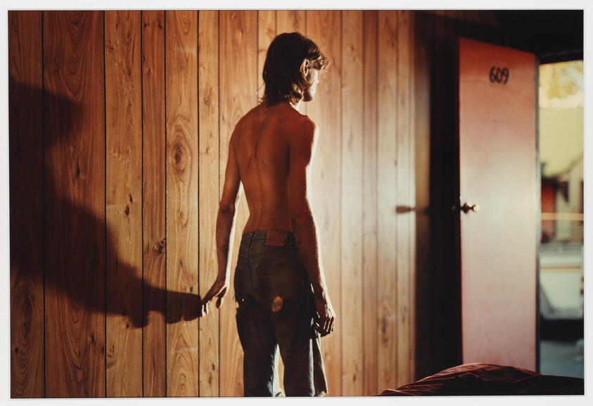 A shirtless young man stands in a wood-paneled room as light casts a long shadow.