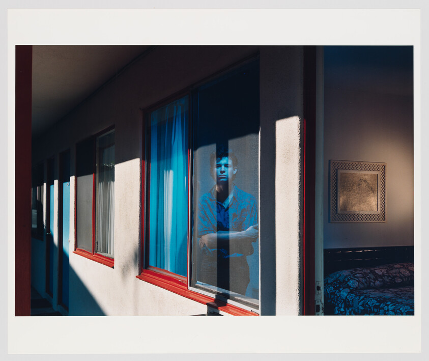 A man stands behind a motel window with arms crossed, bathed in blue light.