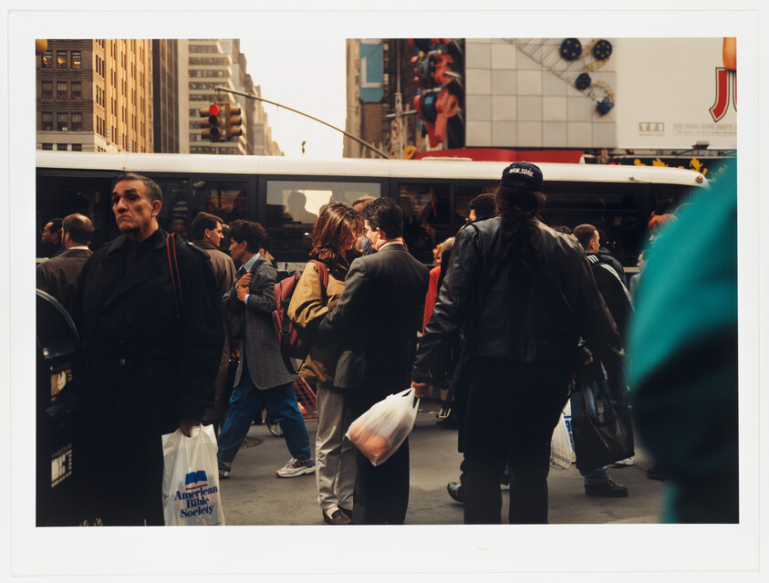 Crowd of commuters wait at a busy city intersection, several people holding shopping bags.