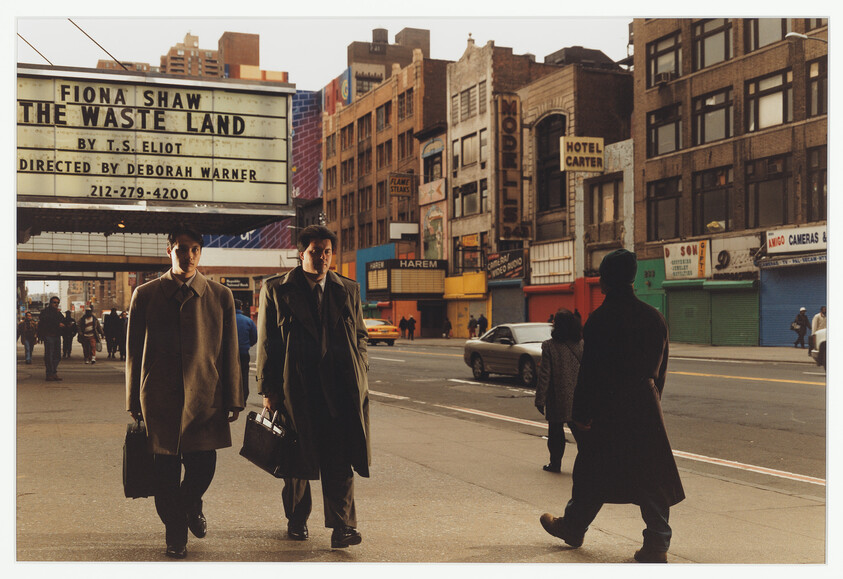 Two men in coats walk past a theater marquee announcing "The Waste Land" on a city street.