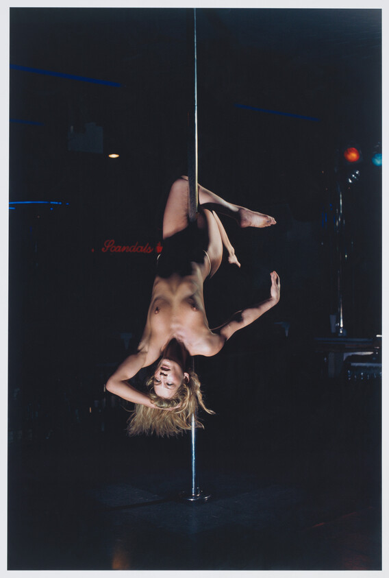 A nude pole dancer hangs upside down from a vertical pole under dim stage lighting.