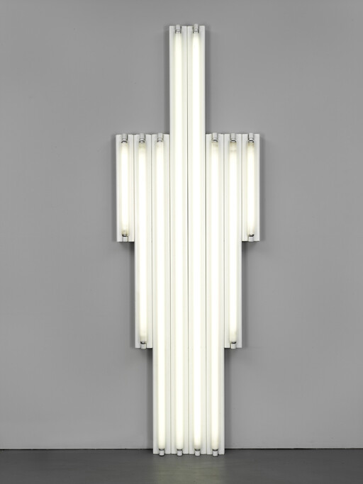 Tall vertical cluster of lit fluorescent tubes mounted on a gray wall in a cross shape.