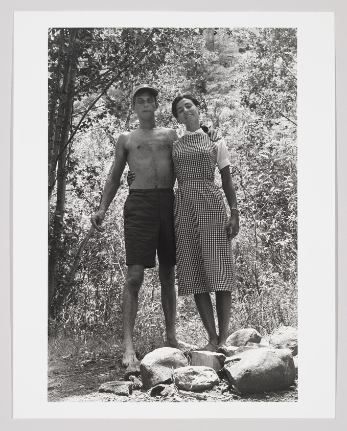 A black and white photograph of a shirtless man and a woman in a gingham dress standing close together on a rocky ground with trees and foliage in the background. The man has his left arm around the woman, and they both have a relaxed posture with a serene expression on their faces.
