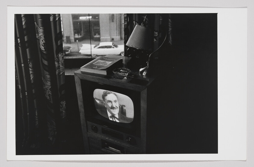 A black and white photo depicting a vintage television set in a dark room, displaying a smiling man in a suit. On top of the TV, there's a desk lamp, a book, and a typewriter. Heavy patterned curtains partially obscure the view of a window that looks out onto a street scene with a parked car.