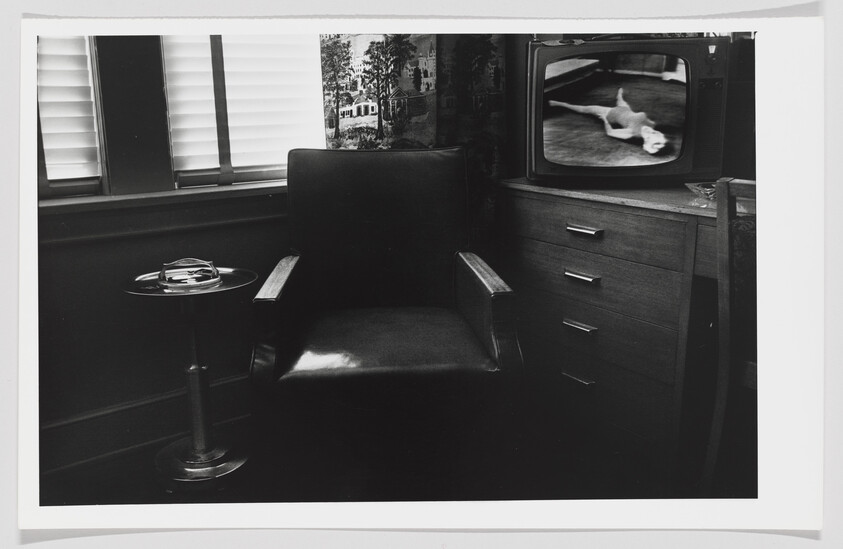 A black and white photograph of a vintage interior scene featuring a leather armchair, a small round table with a glass ashtray, and a wooden television set displaying an image of a cat. The room has a window with closed blinds and a wall adorned with a large framed picture. A wooden chest of drawers stands next to the TV.