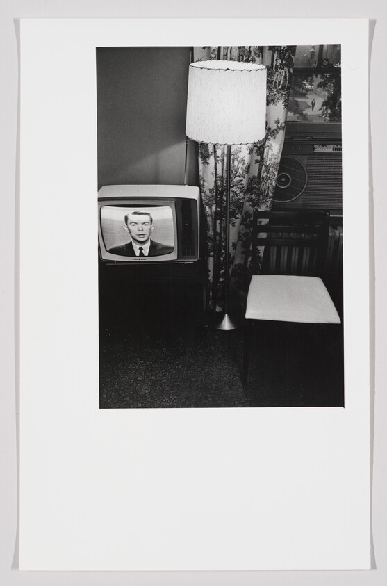 A black and white photograph depicting a vintage television set displaying a man's face, next to a floor lamp with a fringed shade, and a patterned curtain in the background. A wooden chair and a part of an air conditioning unit are also visible. The scene suggests a mid-20th-century living room setting.