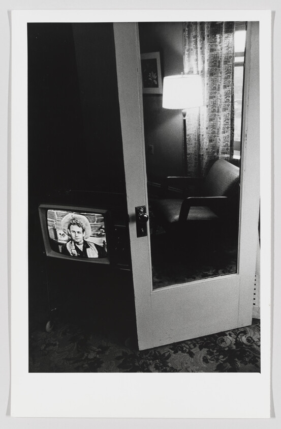 A black and white photograph showing a partially open door leading into a room with a vintage television set on, displaying a person's face. To the right, there's a lamp on a table illuminating patterned curtains beside a window, with a chair visible in the softly lit interior. The carpeted floor extends from the foreground into the room, featuring a floral design.