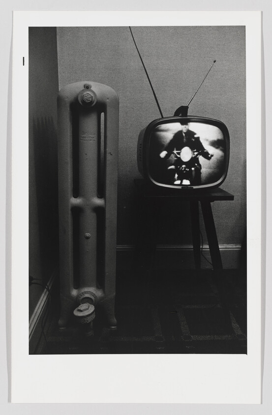 A black and white photograph featuring a vintage television set on a small stand displaying an image of a person riding a motorcycle. To the left of the television is a tall, old-fashioned radiator. The setting appears to be a simple room with patterned carpeting, and the television has rabbit ear antennas.