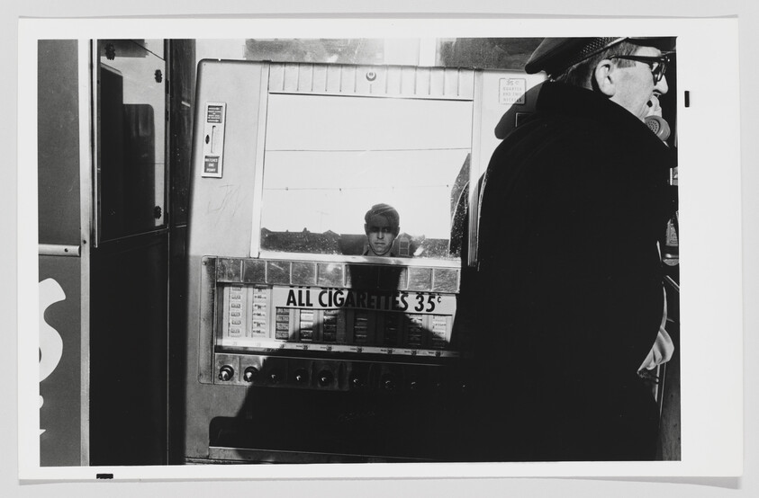 A black and white photograph capturing a moment at a cigarette vending machine. A man in a cap and glasses is seen from the side, possibly making a selection or purchase. Reflected in the glass of the machine is the face of a young person looking directly at the camera. The machine advertises "ALL CIGARETTES 35¢" with various brands and prices listed below. The setting appears to be indoors, with a glimpse of the outside visible in the reflection.