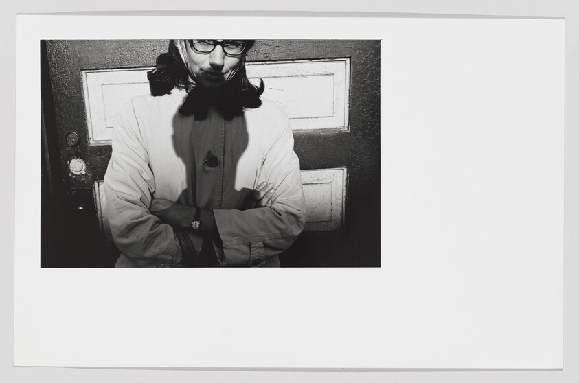 Person with glasses stands in front of a door with arms crossed, shadow across their face.