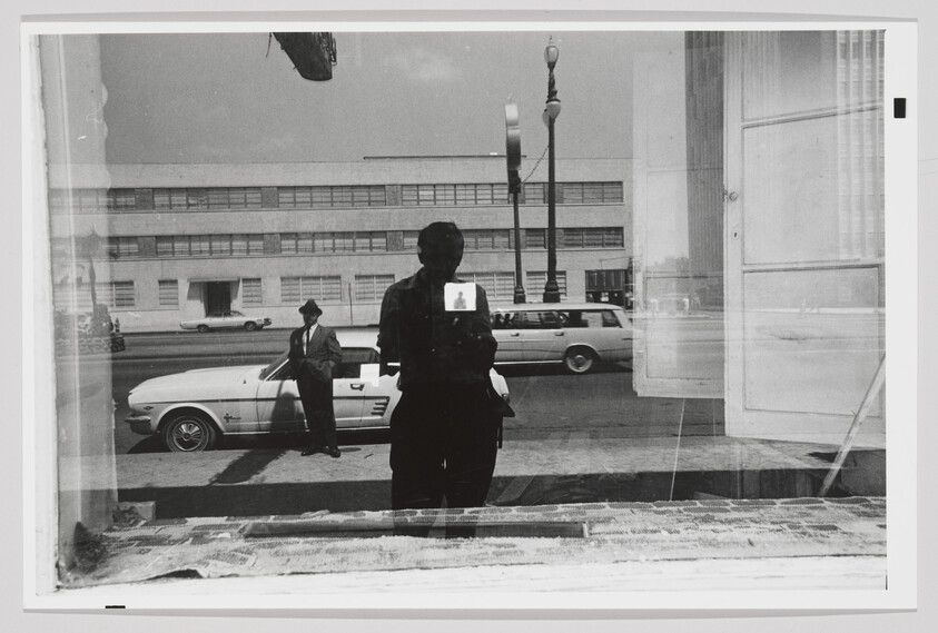 A photographer reflected in a shop window with a man standing by a parked car on the street.