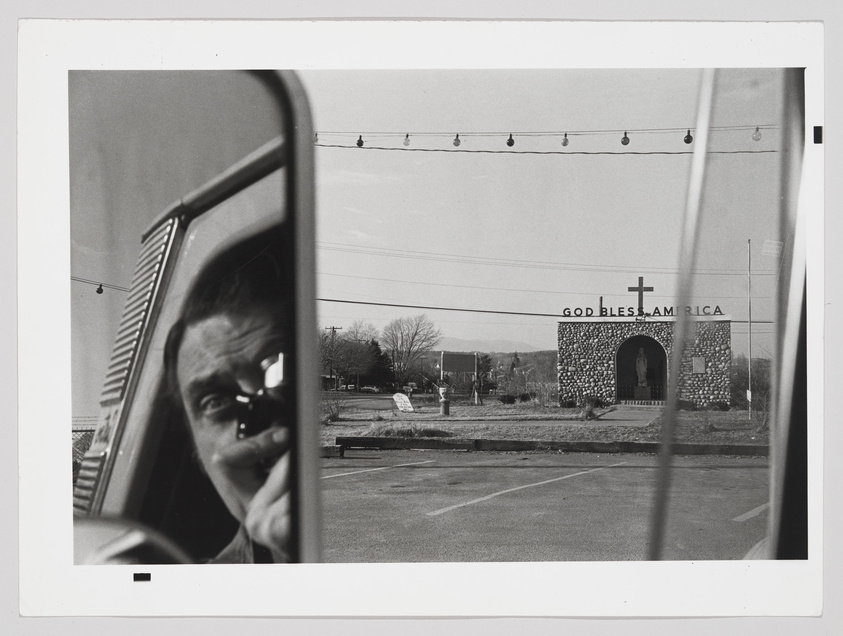 A black and white photograph showing a reflection of a man's face in a car's side mirror, with a structure in the background bearing a cross and the words "GOD BLESS AMERICA" on its facade. Power lines and a clear sky are visible above the structure.