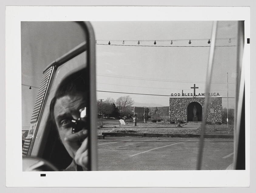 Person taking a photograph reflected in a car mirror with a small stone chapel reading "God Bless America" in background.