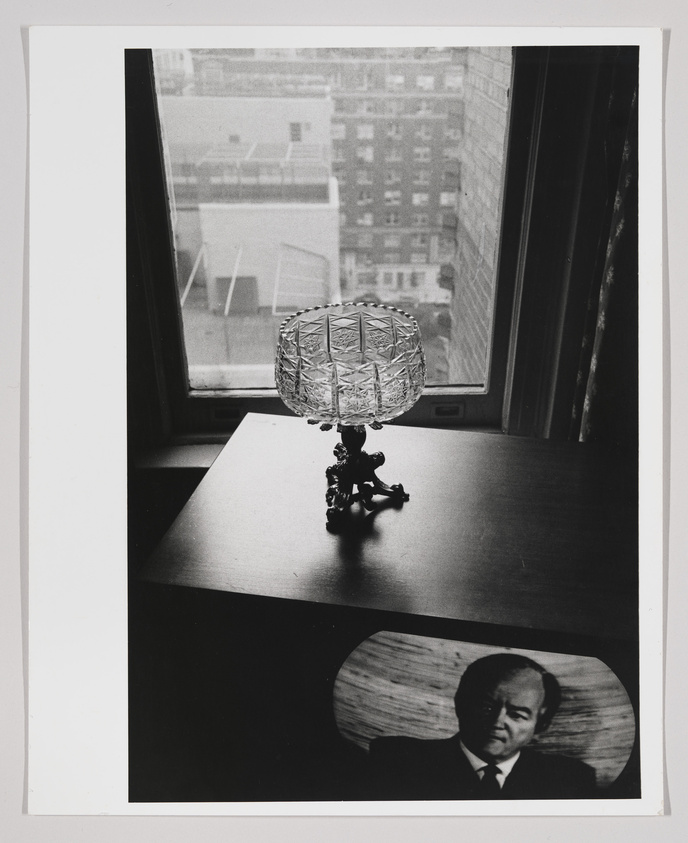 A black and white photograph featuring a crystal table lamp on a wooden desk, with a cityscape visible through a window in the background. In the foreground, there's a magazine or newspaper with a photo of a man's face visible on the desk. The image has a contrast of indoor and outdoor elements, with a focus on the lamp and the printed face.