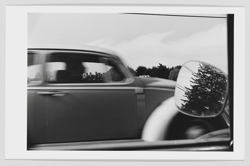 A black and white photograph capturing a motion-blurred car passing by the viewer's car, with a clear reflection of trees in the side mirror.