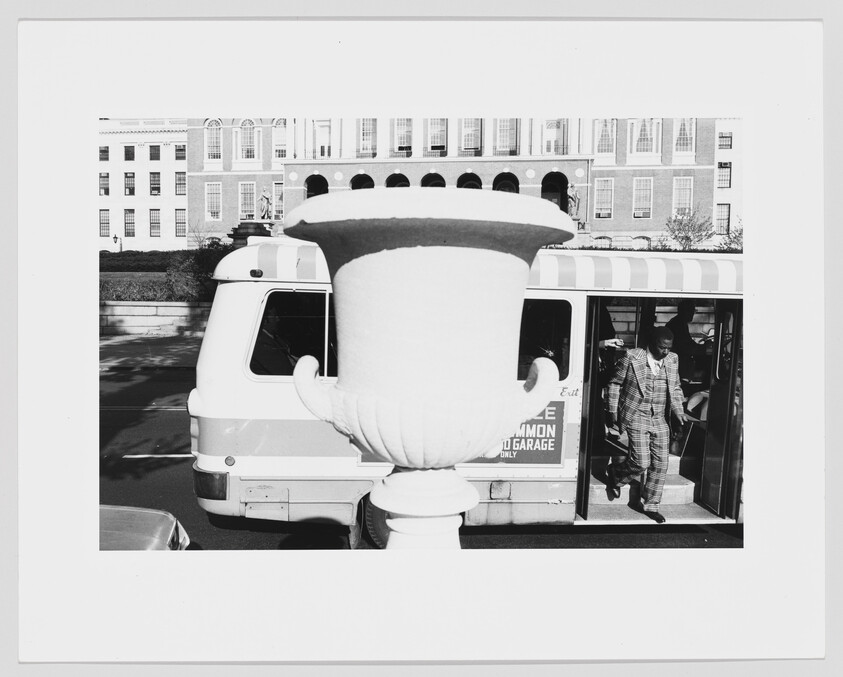 A large decorative urn stands in front of a bus while a man in a plaid suit steps off.
