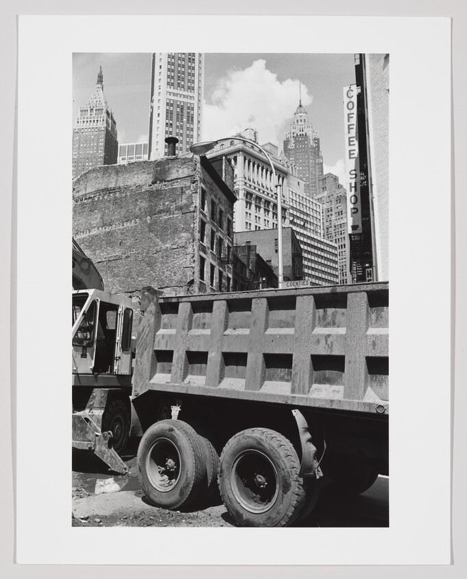 A parked dump truck sits at a construction site with tall city skyscrapers and a coffee shop sign.