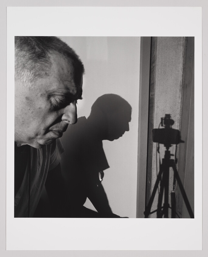 An older man in profile sits near a wall, his shadow and a camera tripod shadow visible.