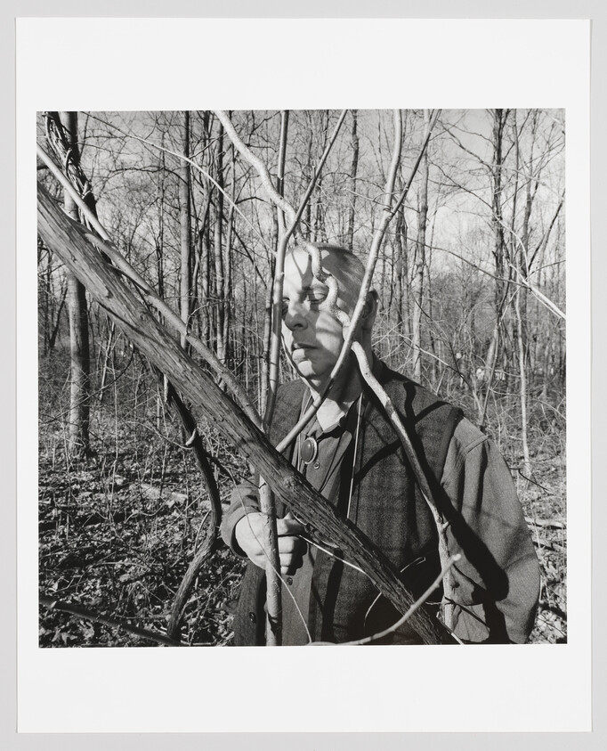 An older man stands among leafless trees with branches crossing his face while holding a walking stick.