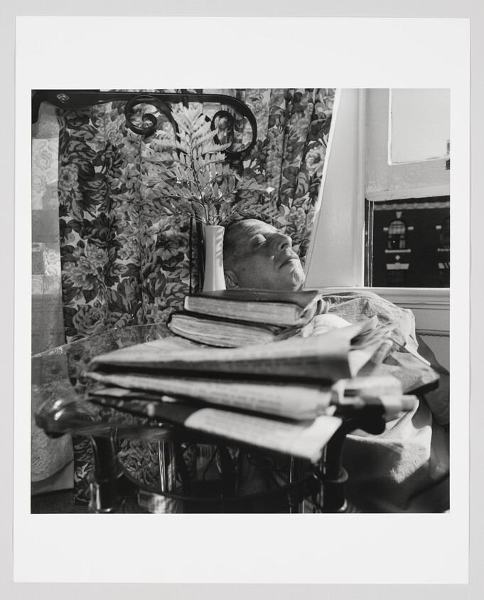 An elderly man sleeps in a chair beside a small table stacked with books and newspapers.