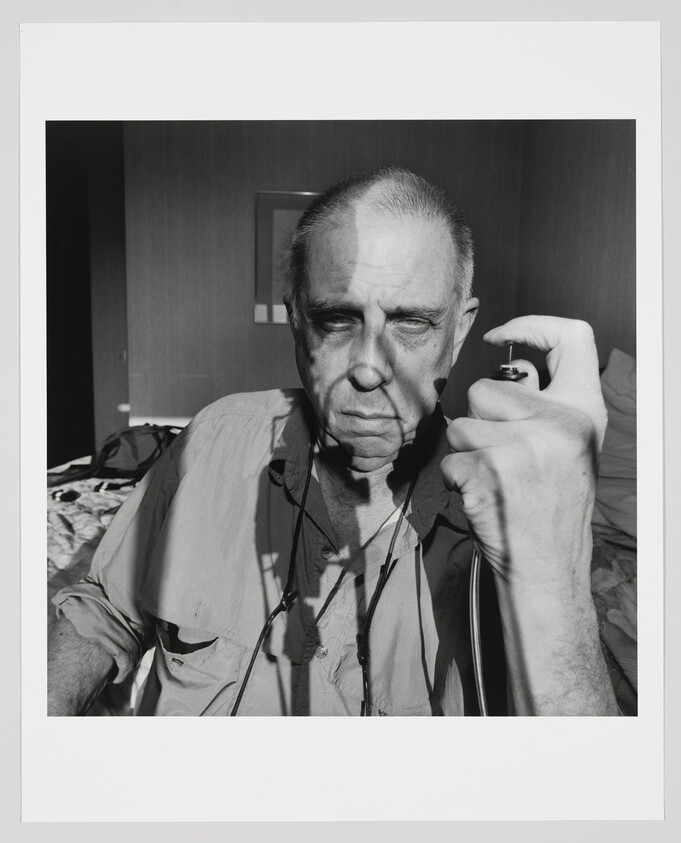 An elderly man sits in a room, gripping a small spray can near his face.
