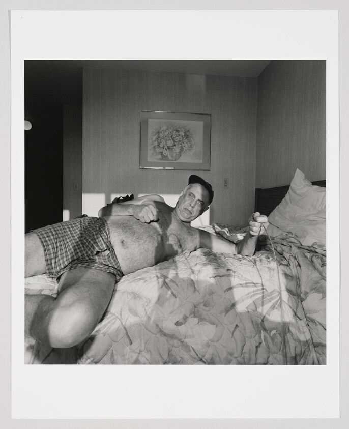 A shirtless man in plaid boxers lies on a bed holding a cord and looking at the camera.