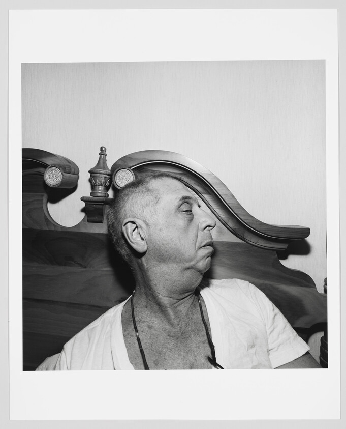 Elderly man sitting up in bed looking to his right against a carved wooden headboard.