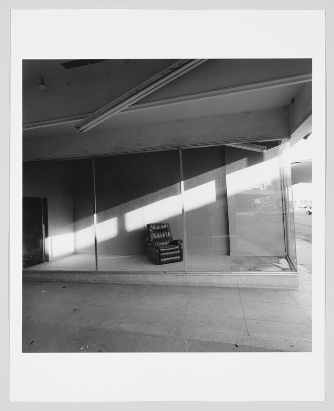 A single worn leather armchair sits alone in an empty storefront window bathed in sunlight.
