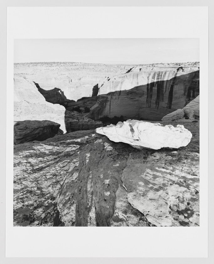 Large flat white rock sits on a layered canyon rim overlooking deep eroded cliffs and valleys.