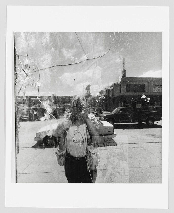 Person taking a photograph reflected in a cracked storefront window with cars passing behind.