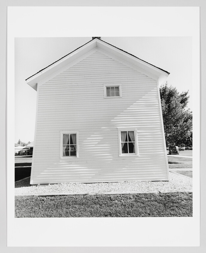 A black and white photograph of a two-story white house with a gabled roof, featuring three visible windows and shadow patterns on the side wall, set against a clear sky with trees in the background. The house is surrounded by a lawn and a gravel area.