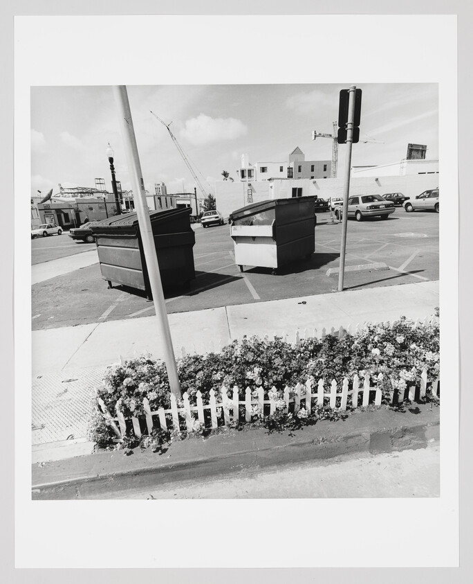 Two large dumpsters sit in a parking lot behind a small white picket fence and flowers.