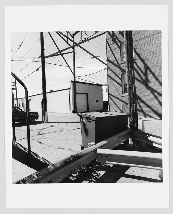 A dumpster sits in an alley beside a brick building under utility poles and wire shadows.