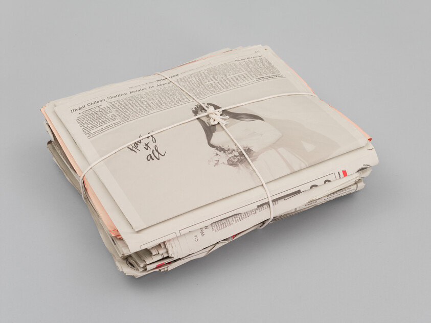 A stack of newspapers bound with string on a plain background, with visible headlines and a partial image of a person on the top paper.