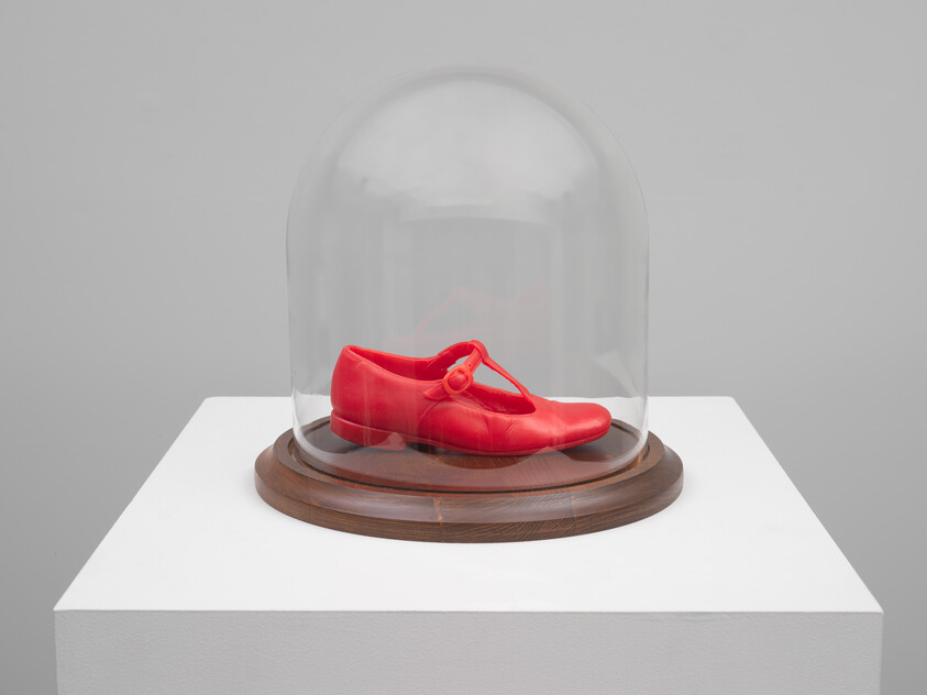 A single bright red ballet shoe is displayed under a clear glass dome, which is placed on a round wooden base. The entire piece is exhibited on a white pedestal against a light grey background.