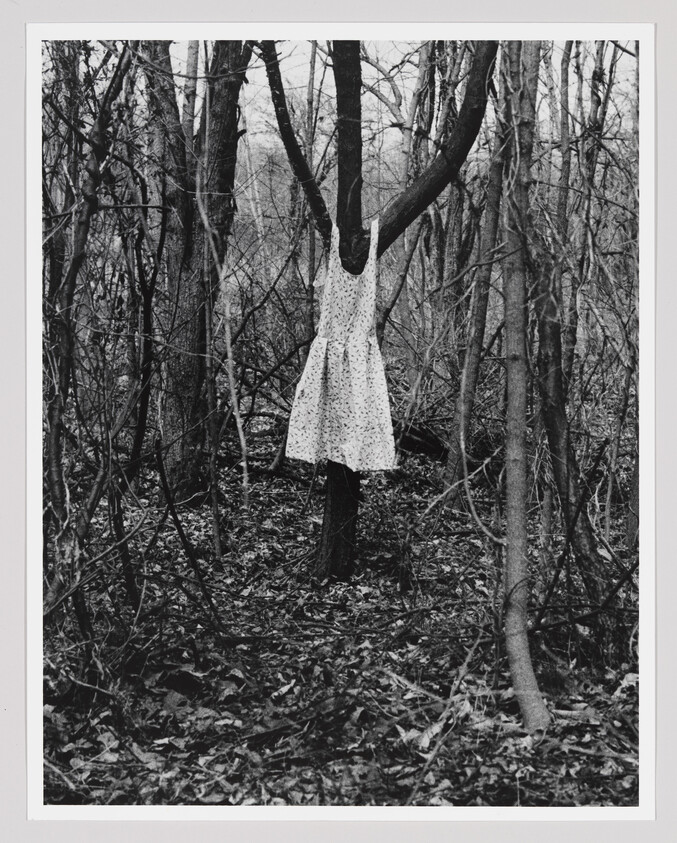 A black and white photograph depicting a leafless forest with a dress on a hanger suspended from a tree branch, creating the illusion of a headless figure standing among the trees. The dress is white with a floral pattern, and the surrounding forest floor is covered with fallen leaves.