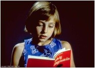 A young girl quietly reading aloud from a small red book against a dark background.