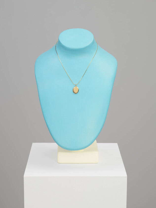 A gold locket necklace displayed on a light blue jewelry bust on a white pedestal.