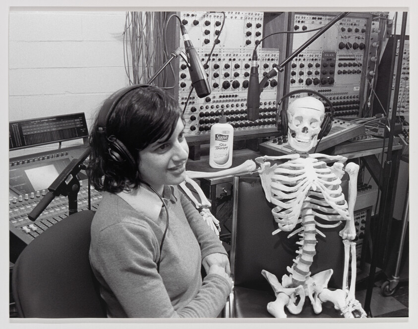 A woman wearing headphones sits smiling beside a skeleton also wearing headphones in a recording studio.