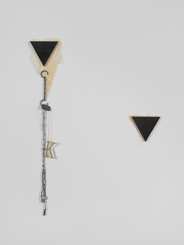 A conceptual art piece featuring two triangular shapes with black tops and beige bottoms, one hanging vertically with decorative elements including a chain, a letter 'K,' and a tassel, and the other smaller triangle positioned separately in the upper right corner against a white background.