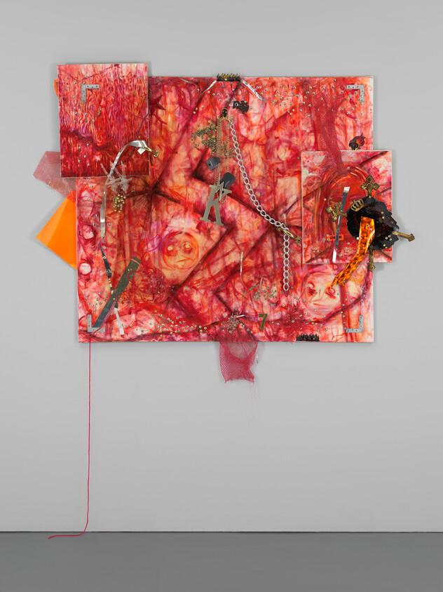 A vibrant mixed media artwork hangs on a white wall, featuring an assemblage of red and pink tones with abstract patterns that resemble floral and organic shapes. Various materials such as chains, beads, and fabric are incorporated into the piece, creating a three-dimensional effect. Some elements extend beyond the main canvas, adding to the dynamic and textured appearance of the artwork. A single red thread dangles towards the floor, drawing the eye downward and emphasizing the piece's layered complexity.