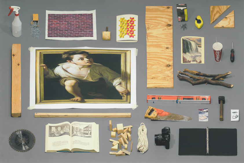 A collage of various objects laid out on a grey surface, including a framed painting of a boy looking outwards, a spray bottle, a brick wall print, a cheese grater with nails, a wooden plank, a level, a handsaw, branches, a circular saw blade, an open book with illustrations, wooden blocks, a coil of rope, a camera, a black folder, and a few other miscellaneous items like a tape measure, a hammer, and a soft drink cup with a straw.