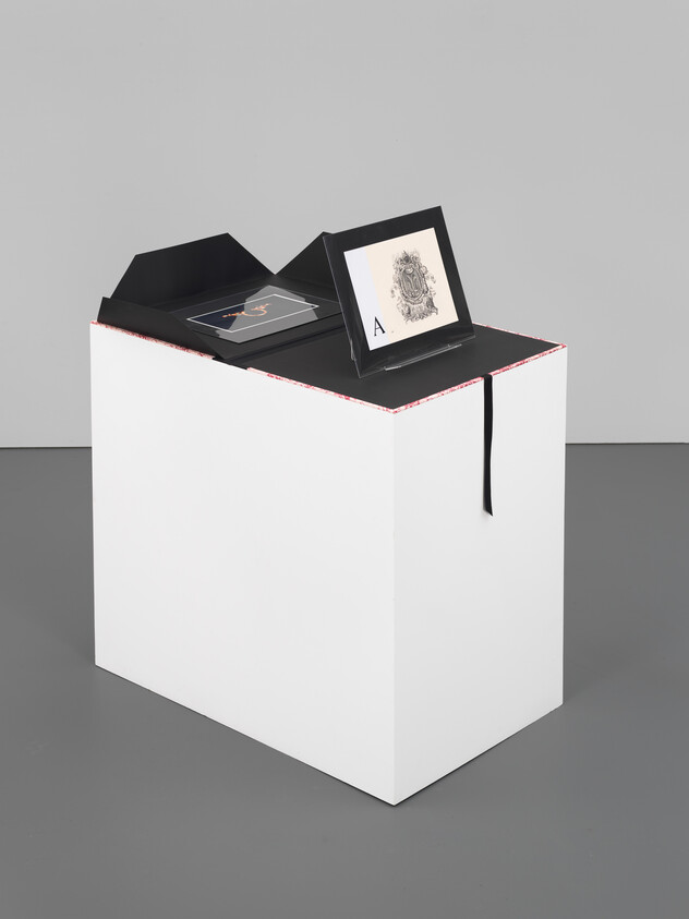 A white display pedestal holds an open black portfolio showing two framed prints and a ribbon.