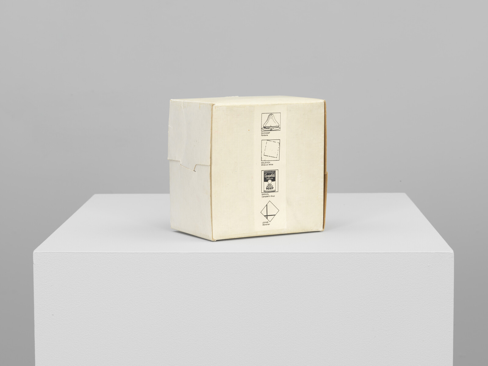 Beige cardboard box with small printed icons sits centered on a white pedestal against gray wall.