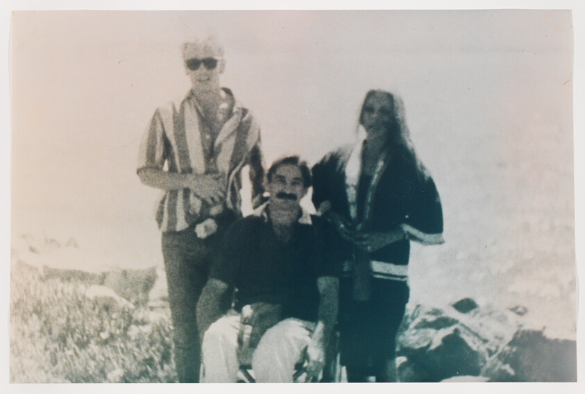 Man in a wheelchair sits outdoors on rocks while two companions stand behind him.