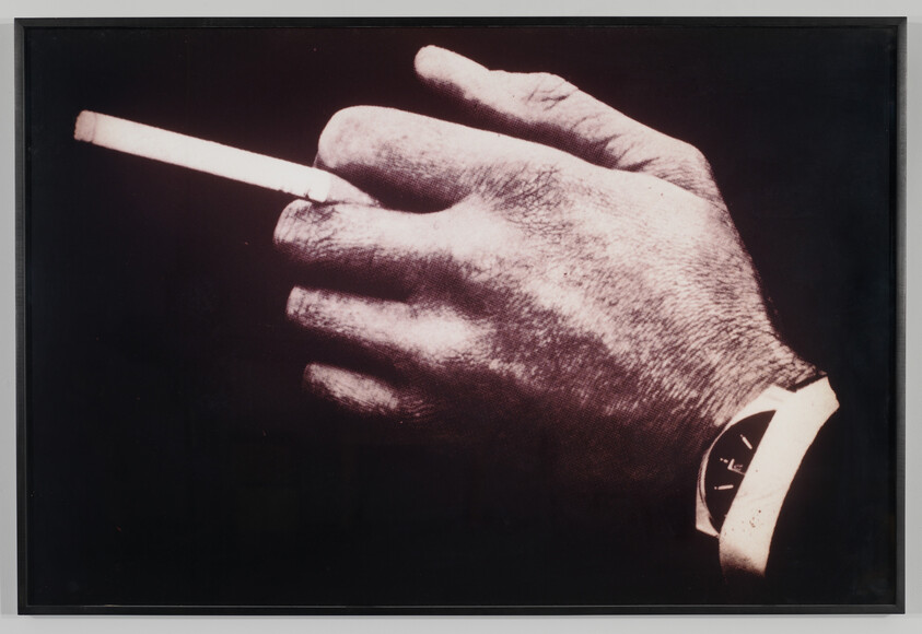 A close-up monochrome photograph of a person's hand holding a cigarette, with a wristwatch visible on the wrist.